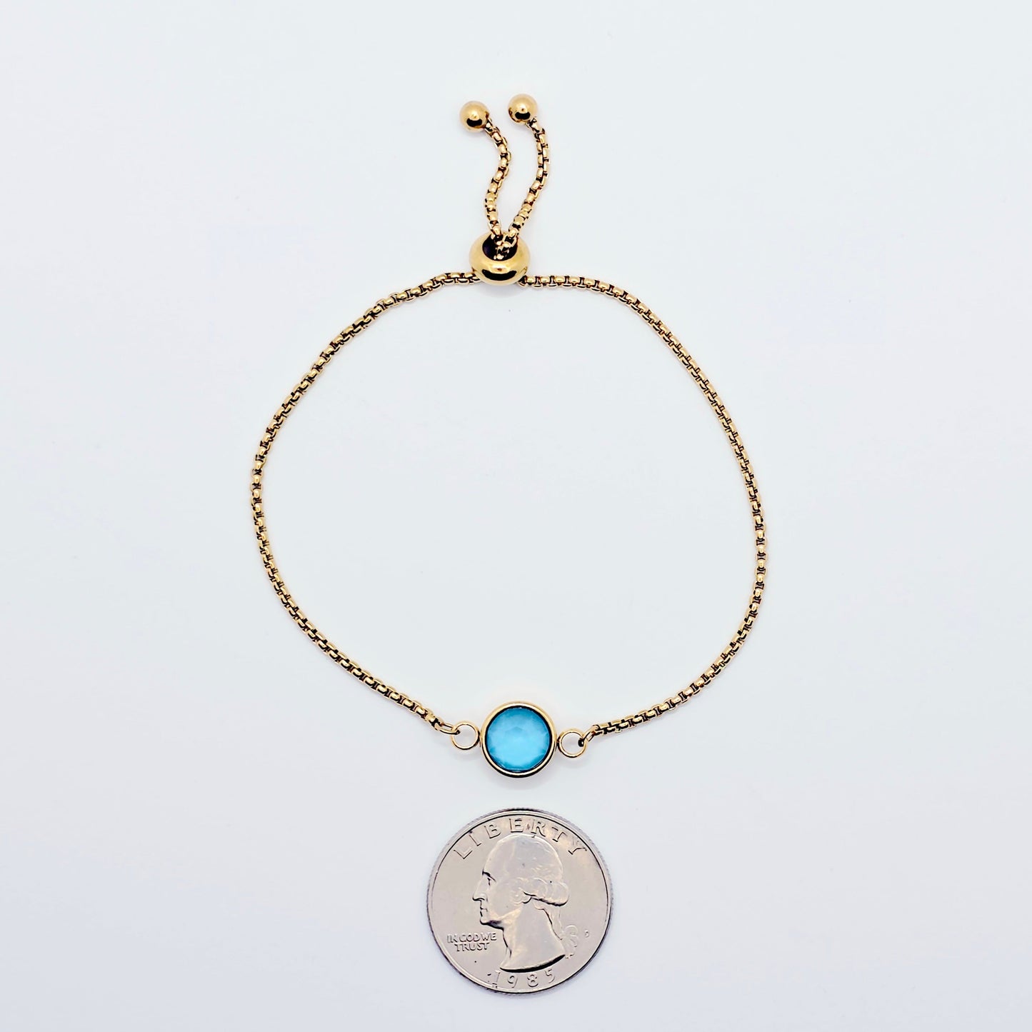 Gold penis bracelet with a blue gemstone next to a quarter for size comparison on a white background