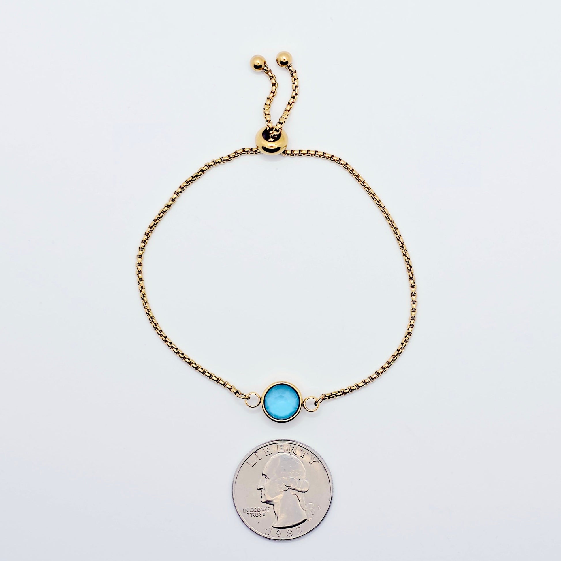 Gold penis bracelet with a blue gemstone next to a quarter for size comparison on a white background