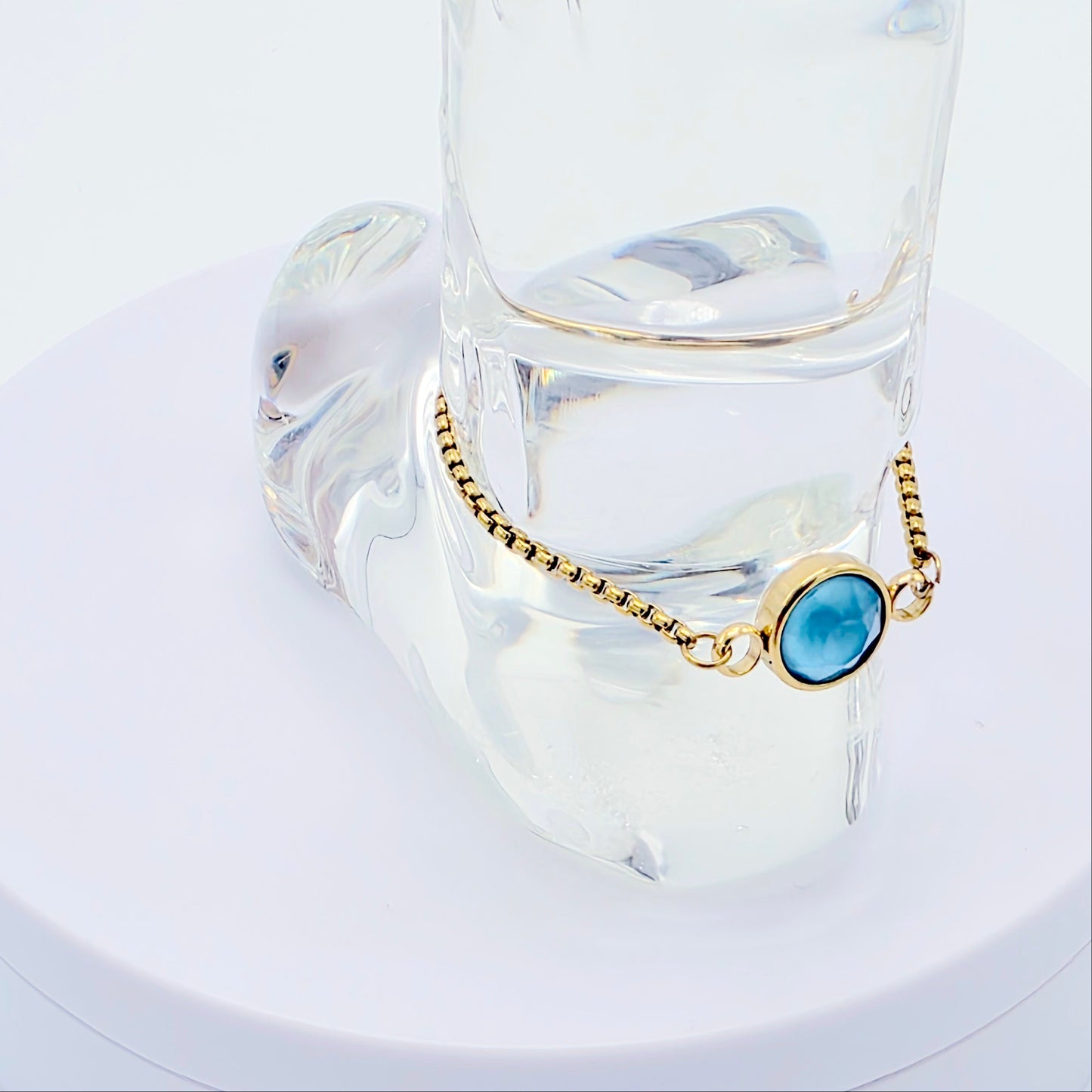 Gold penis bracelet with a blue gemstone on a clear glass stand against a white background