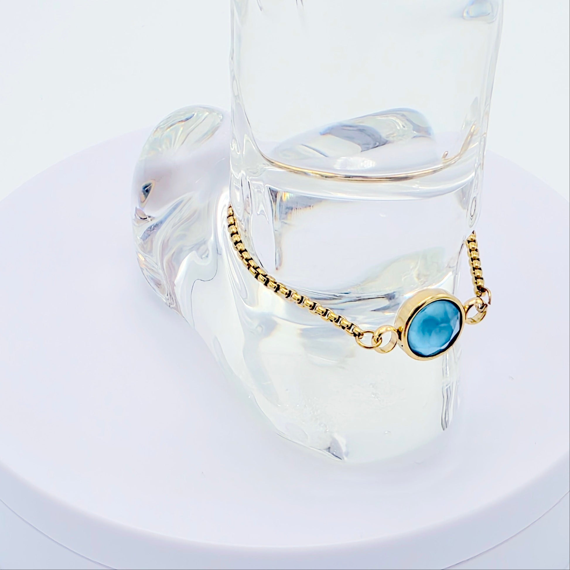 Gold penis bracelet with a blue gemstone on a clear glass stand against a white background