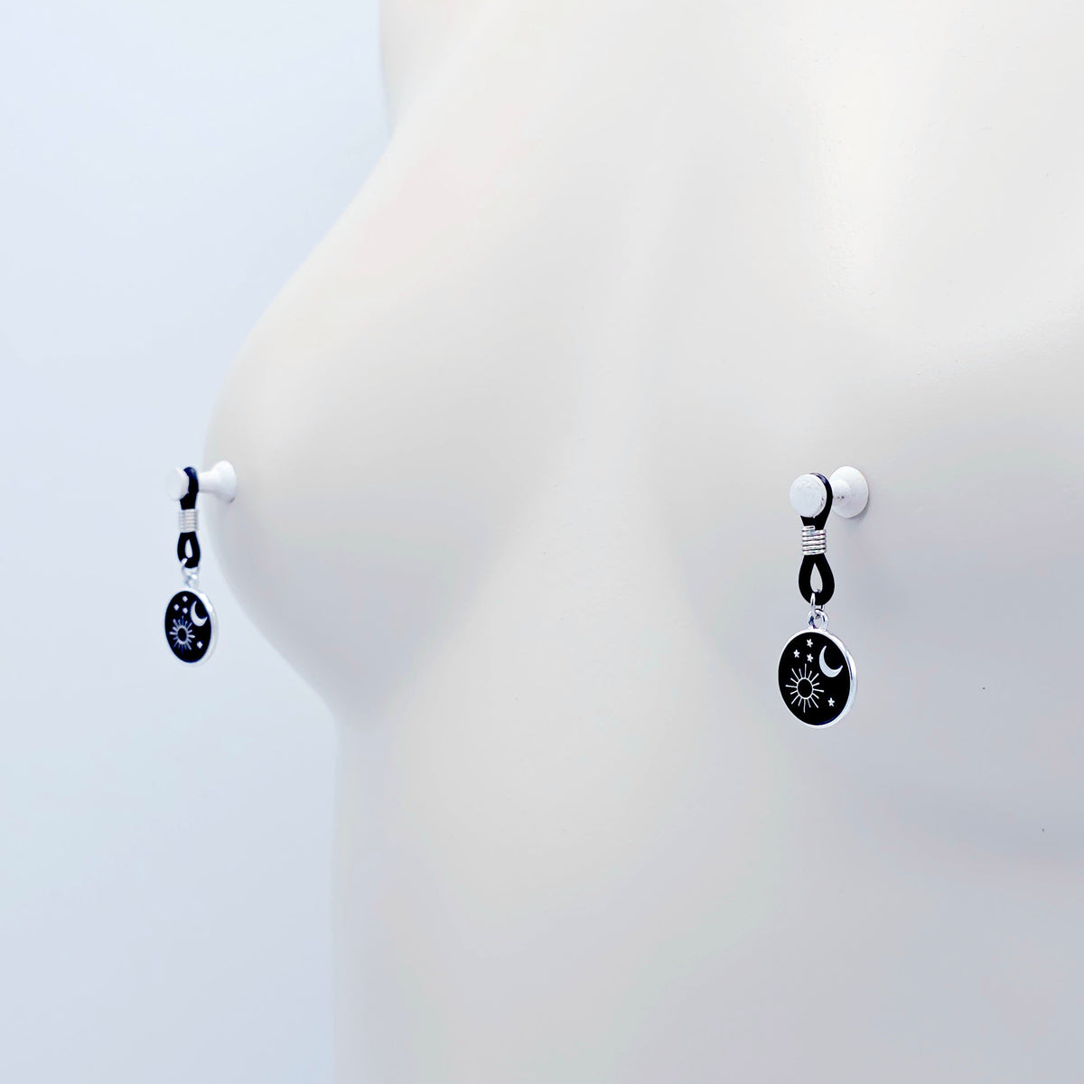 pair of small black nipple nooses with circular celestial charms featuring a crescent moon, sun, and stars on a light gray background.