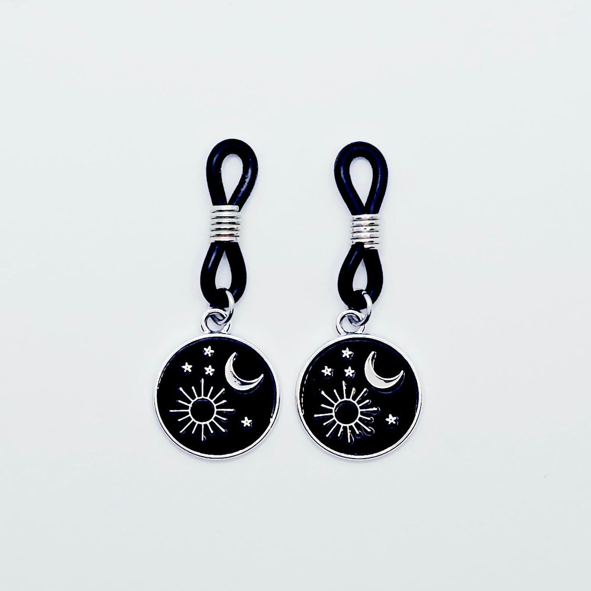 pair of small black nipple nooses with circular celestial charms featuring a crescent moon, sun, and stars on a light gray background.