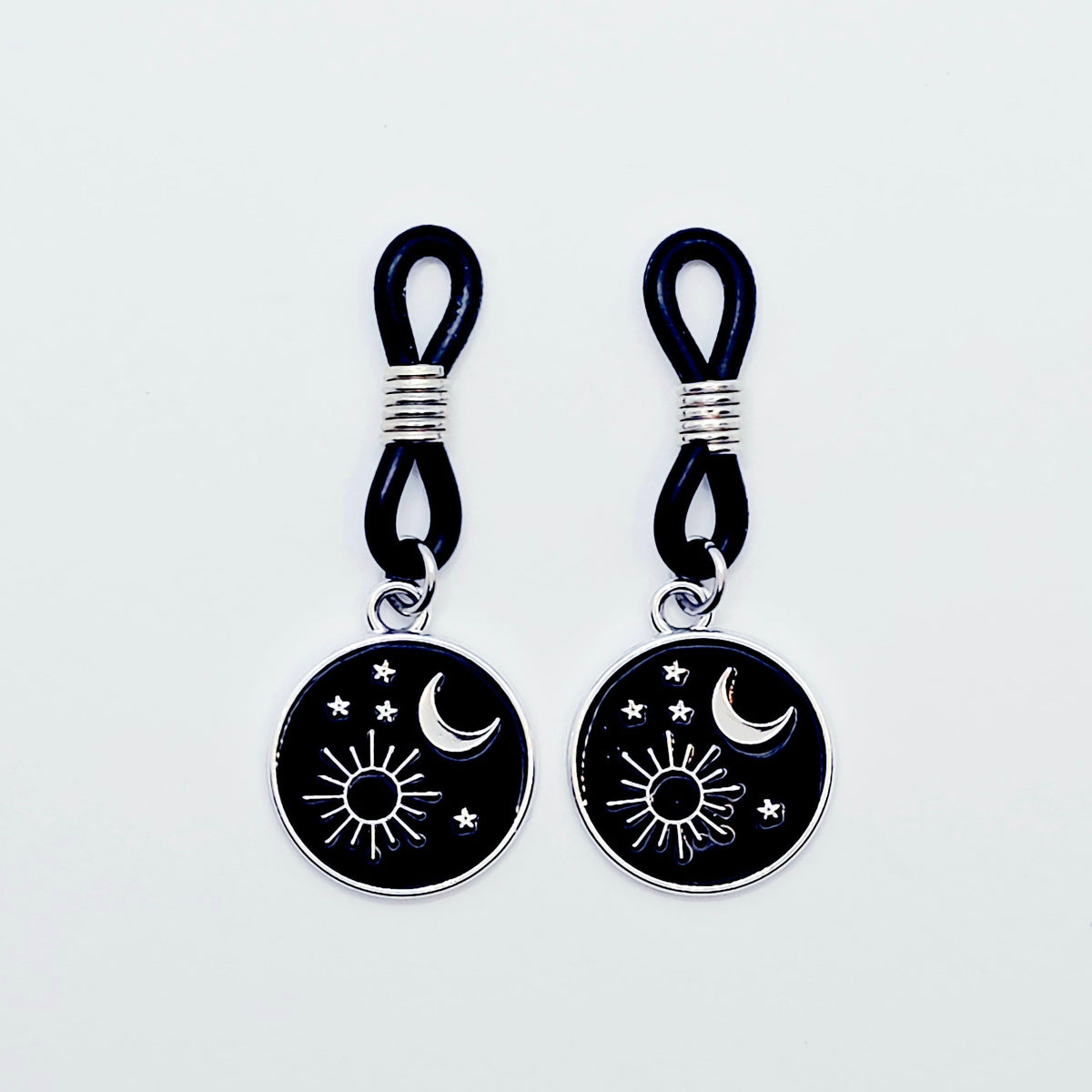 pair of small black nipple nooses with circular celestial charms featuring a crescent moon, sun, and stars on a light gray background.
