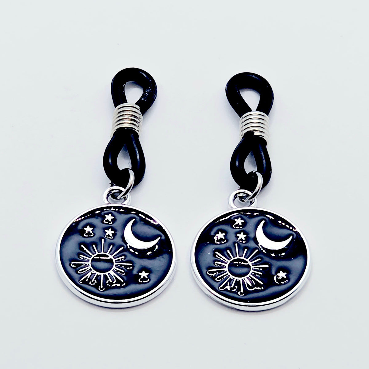 pair of small black nipple nooses with circular celestial charms featuring a crescent moon, sun, and stars on a light gray background.
