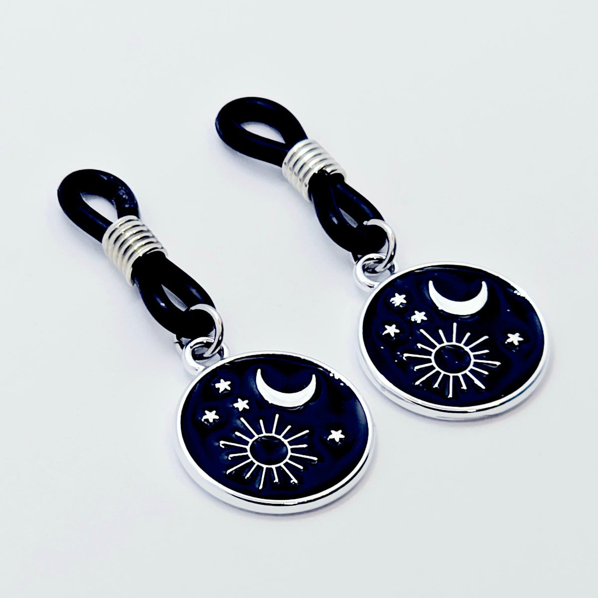 pair of small black nipple nooses with circular celestial charms featuring a crescent moon, sun, and stars on a light gray background.