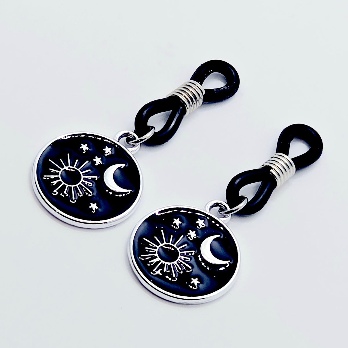 pair of small black nipple nooses with circular celestial charms featuring a crescent moon, sun, and stars on a light gray background.