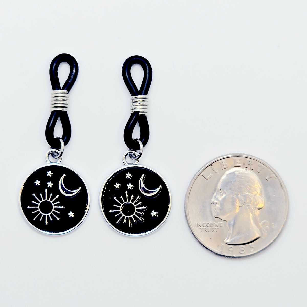 pair of small black nipple nooses with circular celestial charms featuring a crescent moon, sun, and stars on a light gray background.