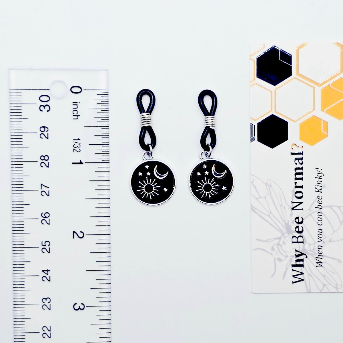 pair of small black nipple nooses with circular celestial charms featuring a crescent moon, sun, and stars on a light gray background.