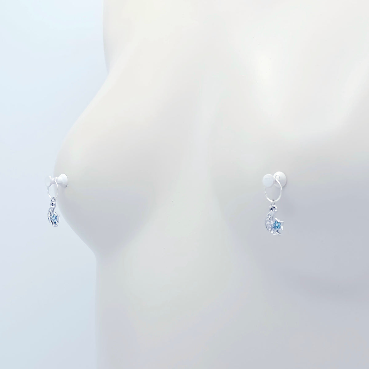 A pair of silver crescent moon and star dangle nipple rings with clear rhinestones and light blue star accents, shown on a mannequin