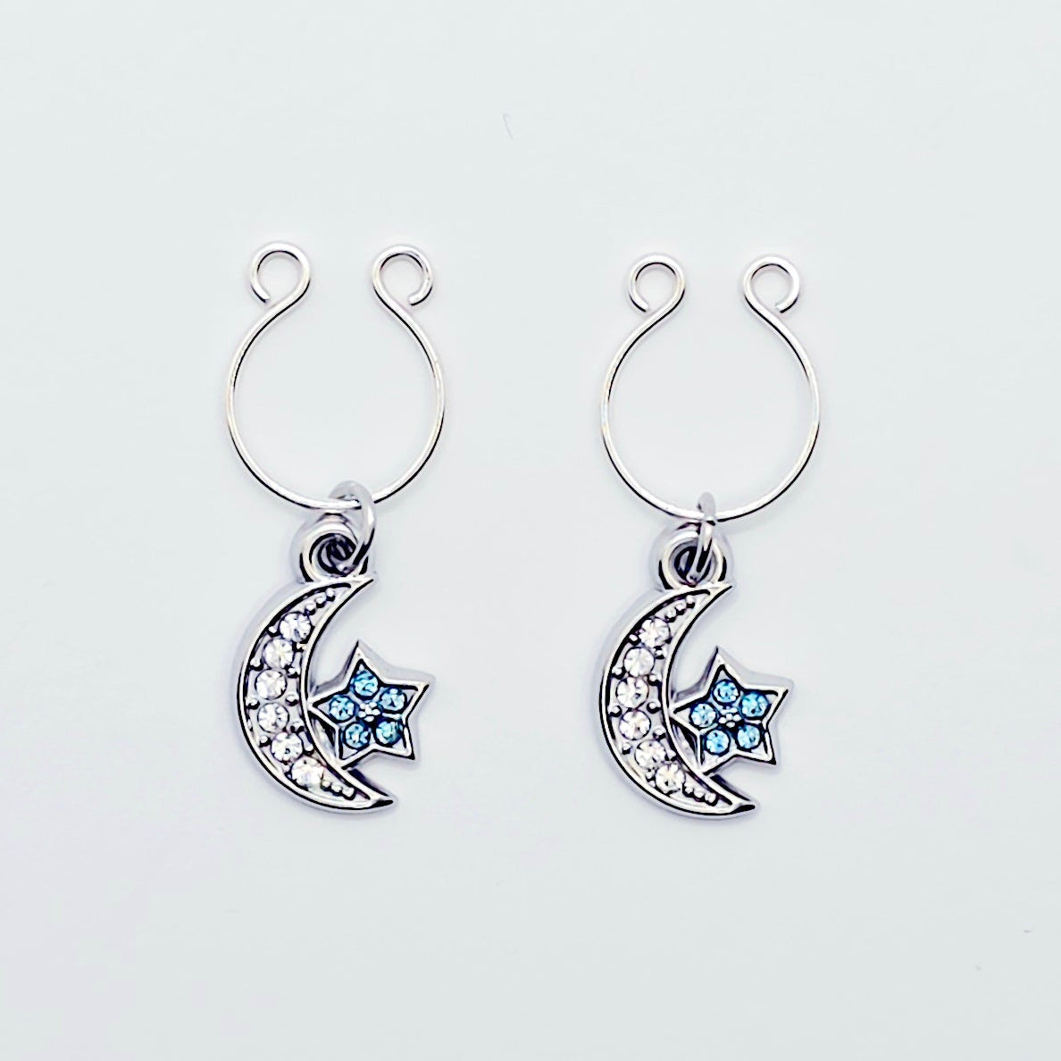 A pair of silver crescent moon and star dangle nipple rings with clear rhinestones and light blue star accents, shown on a plain light gray background.