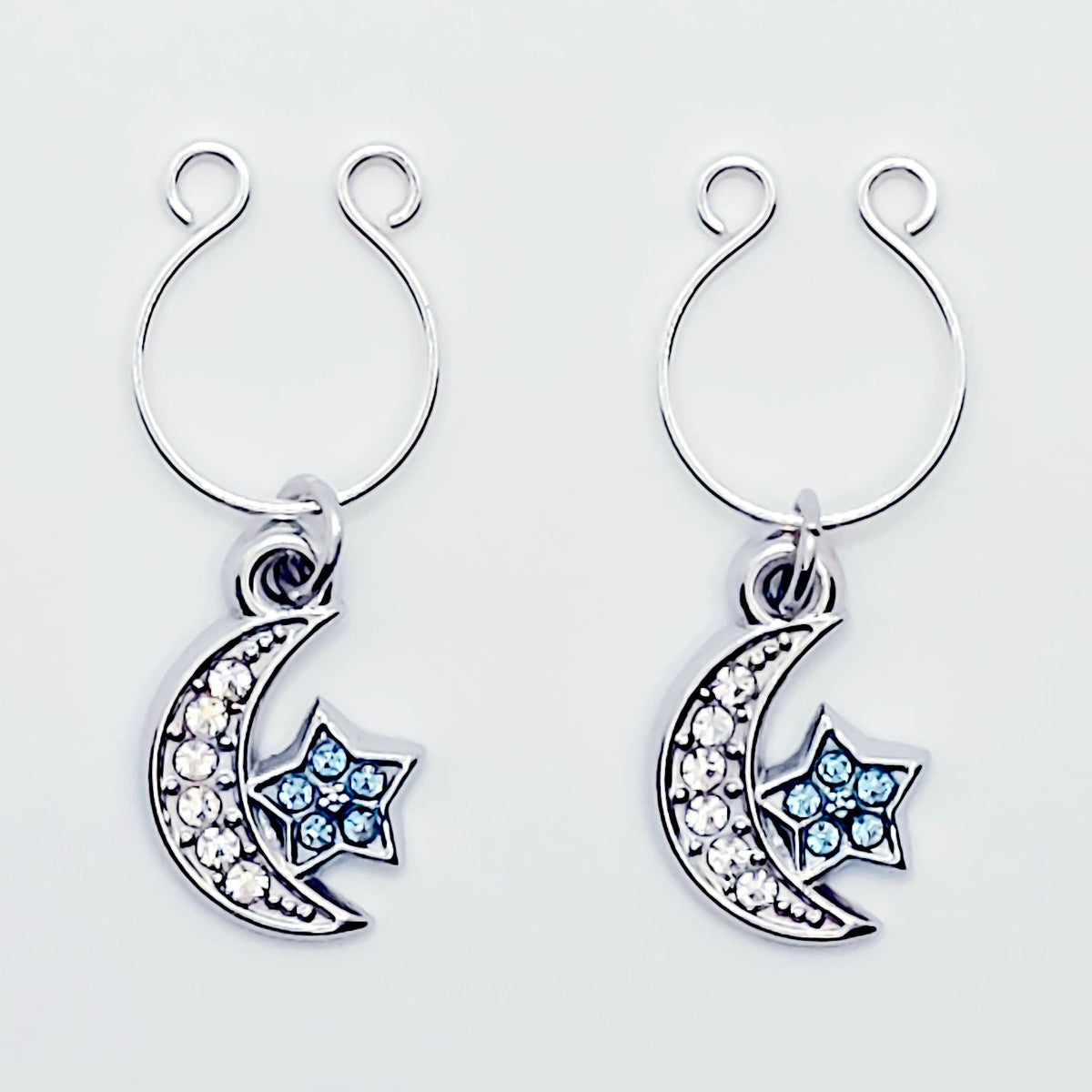 A pair of silver crescent moon and star dangle nipple rings with clear rhinestones and light blue star accents, shown on a plain light gray background.