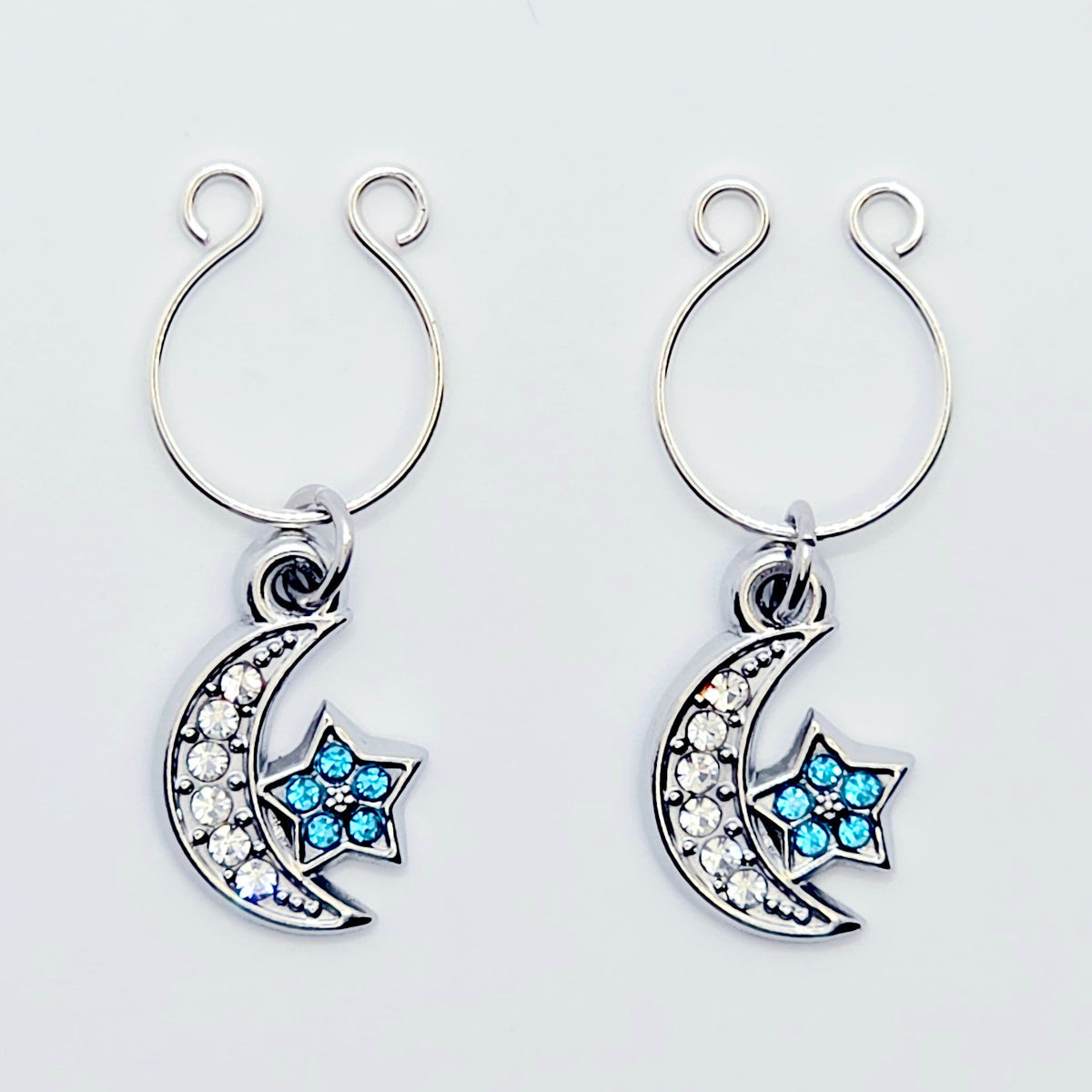 A pair of silver crescent moon and star dangle nipple rings with clear rhinestones and light blue star accents, shown on a plain light gray background.