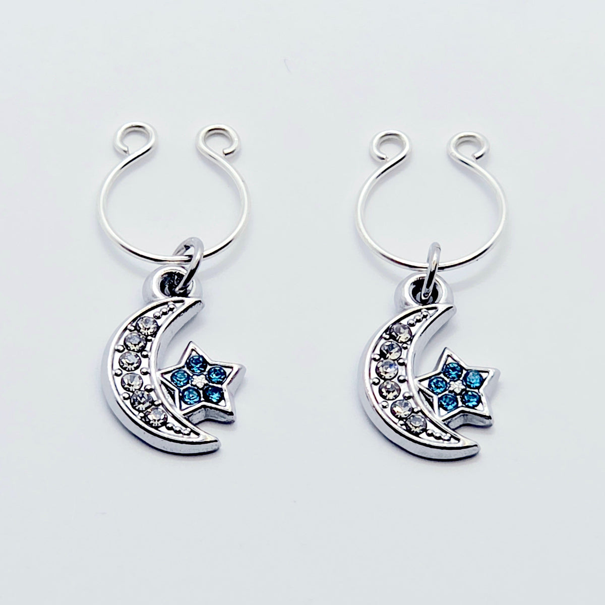 A pair of silver crescent moon and star dangle nipple rings with clear rhinestones and light blue star accents, shown on a plain light gray background.