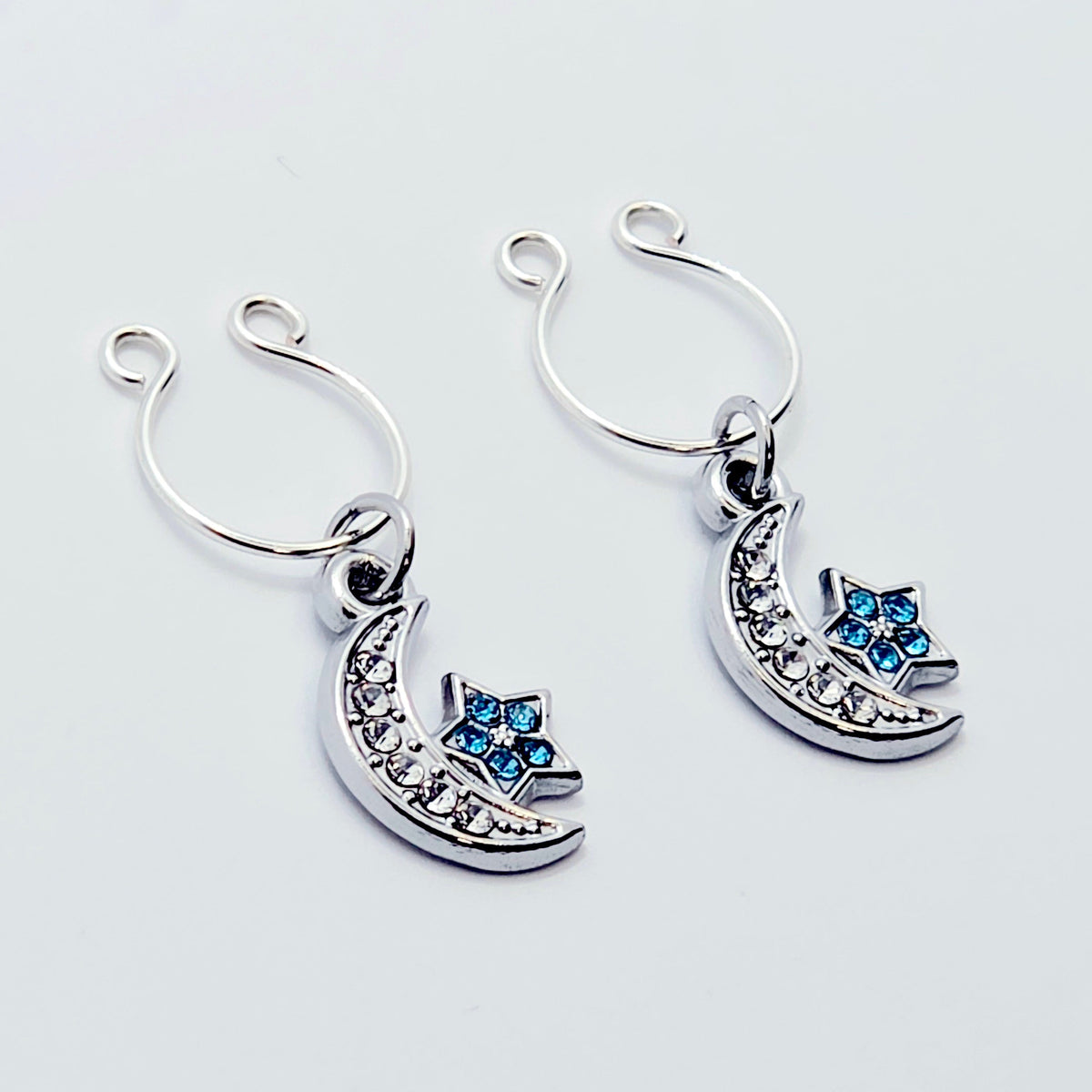 A pair of silver crescent moon and star dangle nipple rings with clear rhinestones and light blue star accents, shown on a plain light gray background.