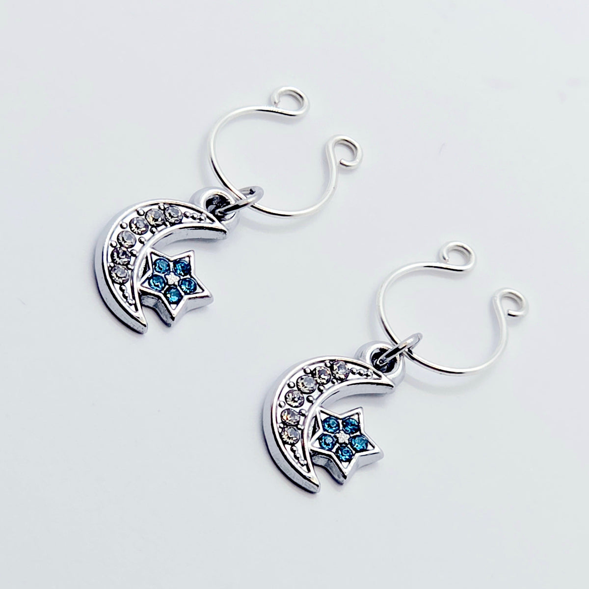 A pair of silver crescent moon and star dangle nipple rings with clear rhinestones and light blue star accents, shown on a plain light gray background.