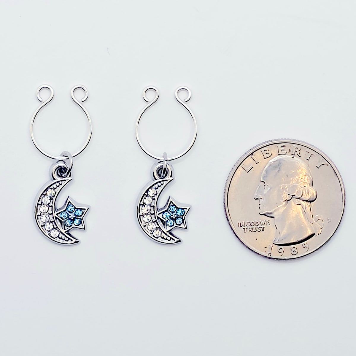 A pair of silver crescent moon and star dangle nipple rings with clear rhinestones and light blue star accents, shown on a plain light gray background. With a quarter t show size. 