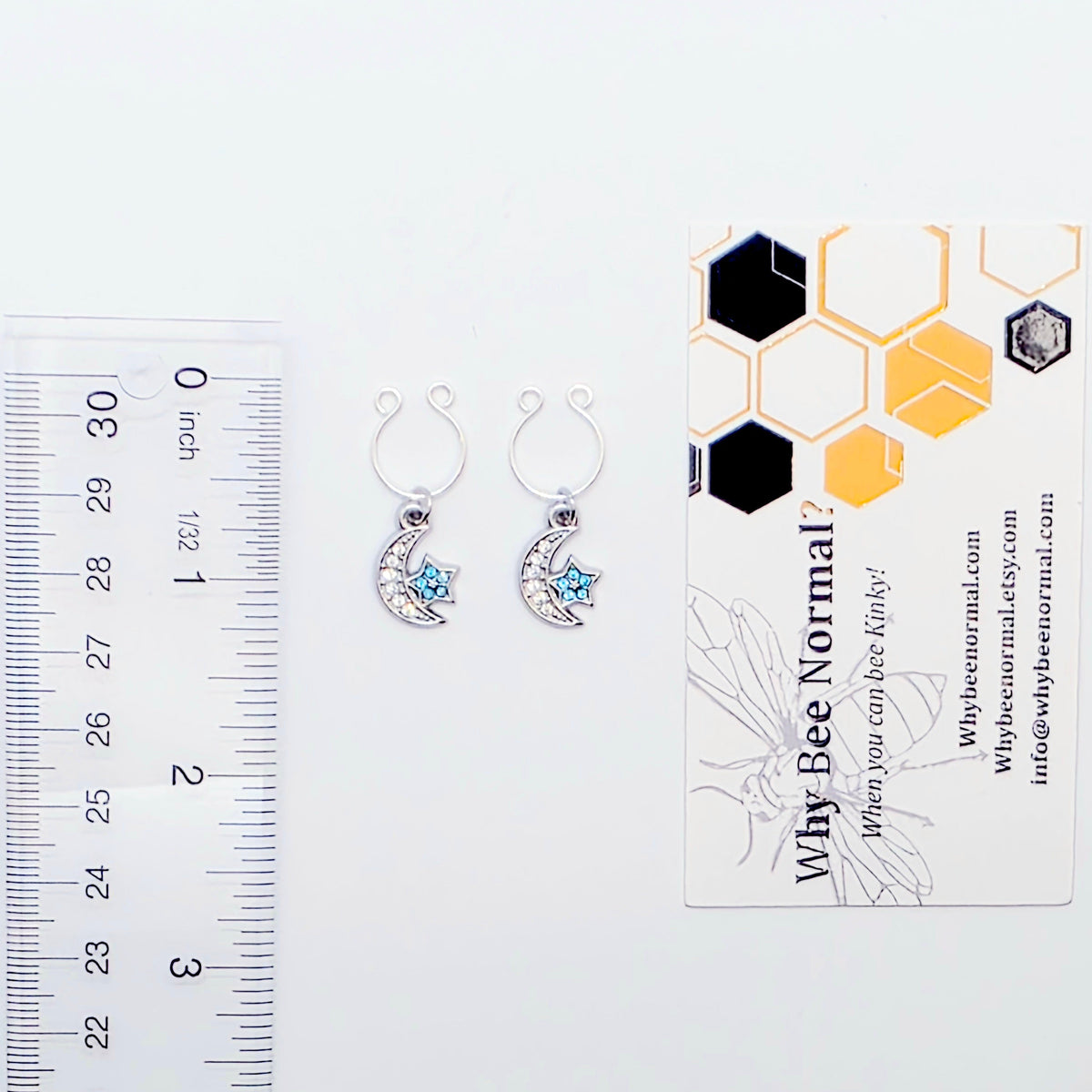 A pair of silver crescent moon and star dangle nipple rings with clear rhinestones and light blue star accents, shown on a plain light gray background with business card and ruler to show size. 