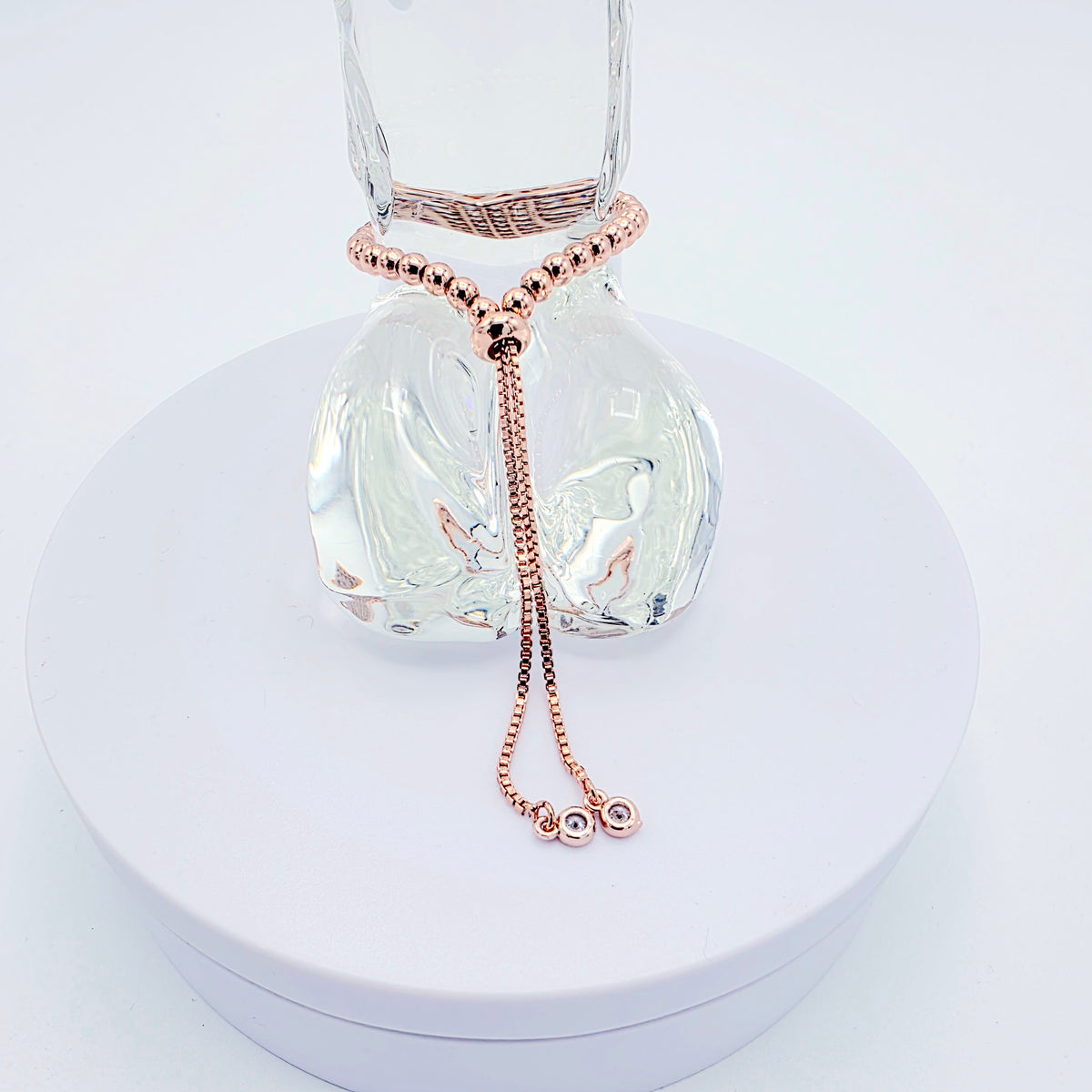 A rose gold beaded bracelet with a long drop chain and small bead charms, displayed on a clear glass bottle over a round white pedestal.