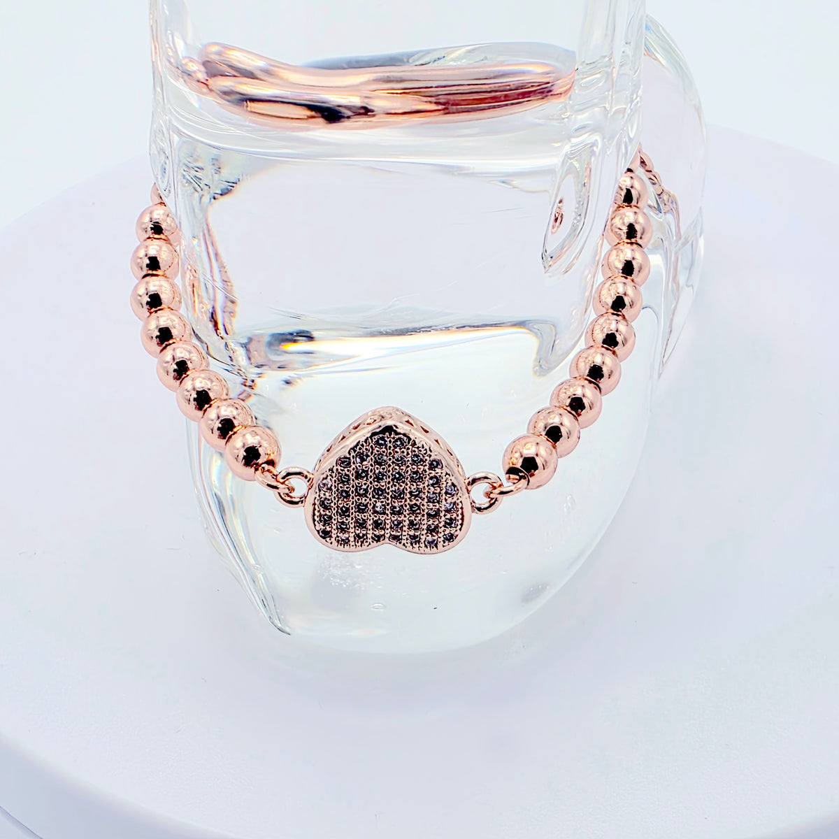 Rose gold beaded necklace with a heart-shaped rhinestone pendant, displayed on a clear glass stand against a white background.