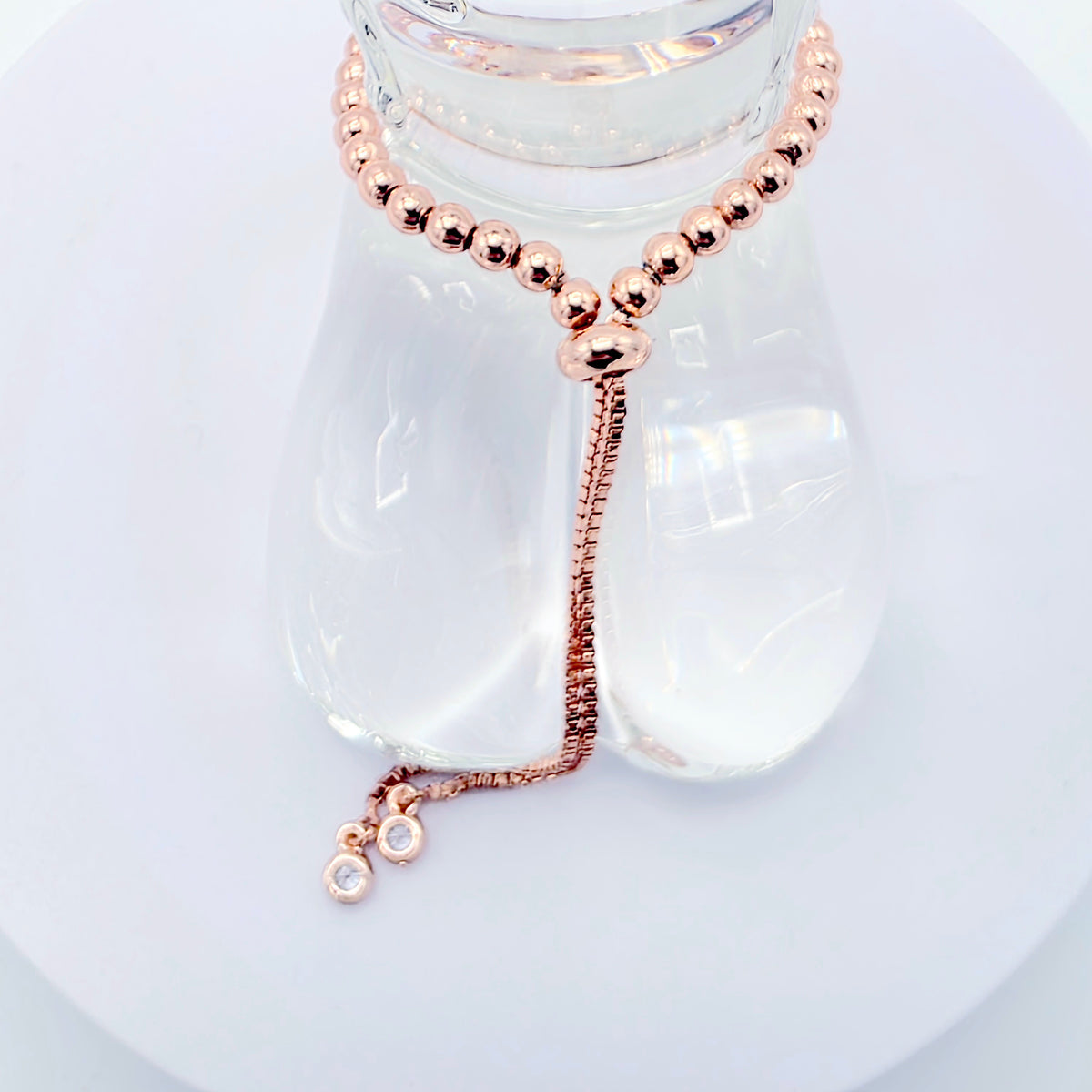 A rose gold beaded choker necklace with a matching adjustable chain and small dangling charm, displayed on a clear glass bottle stand against a white background.