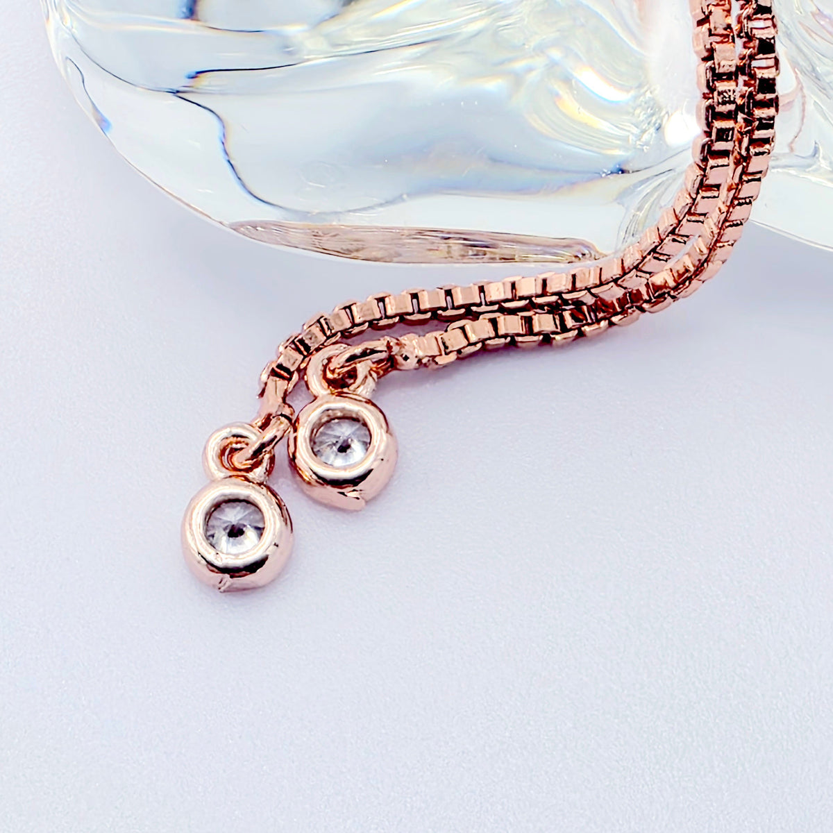 Rose gold chain necklace with two small round bezel-set clear gemstone pendants, displayed on a light gray surface with a translucent glass prop.