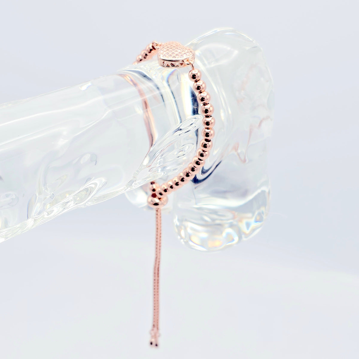 A rose gold beaded bracelet with a matching extender chain, displayed on a clear glass mannequin hand against a white background.