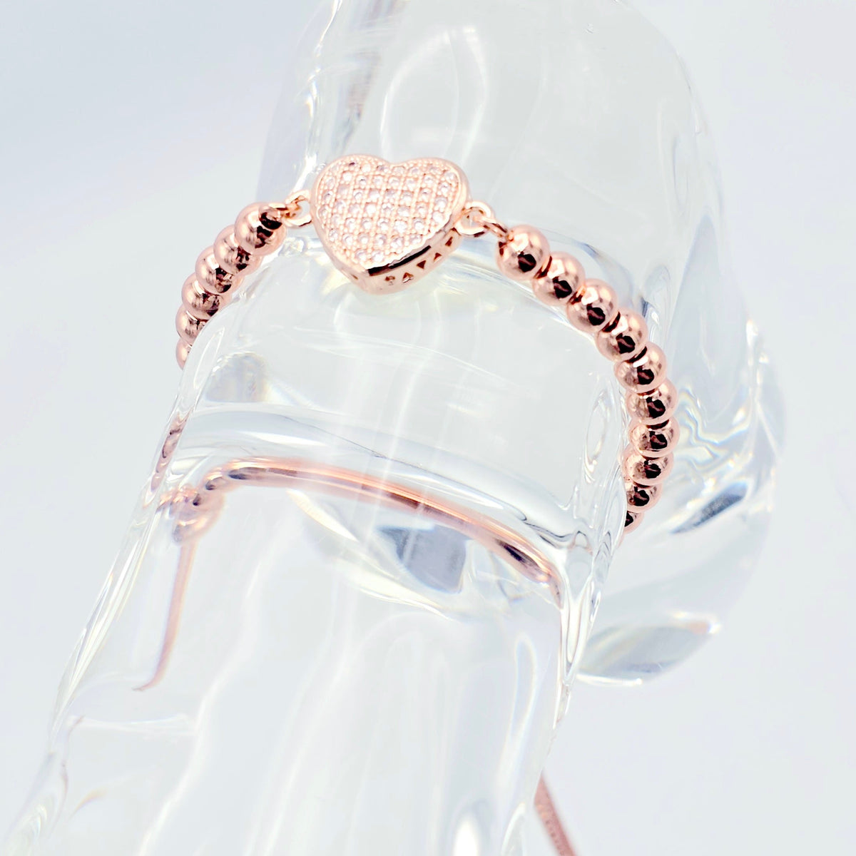 Rose gold beaded bracelet with a textured heart charm, displayed on a clear acrylic jewelry stand against a white background.