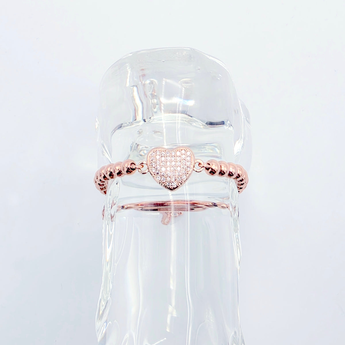 A rose gold heart-shaped ring with a pavé center and textured beaded sides, displayed on a clear glass bottle against a white background.
